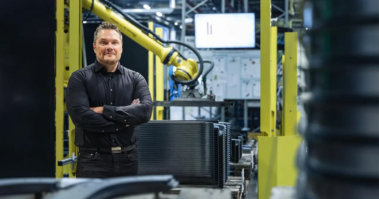 Eine Person steht neben gestapelten Blechkomponenten in einer Fertigungszelle; im Hintergrund ist ein Industrieroboter zu sehen.