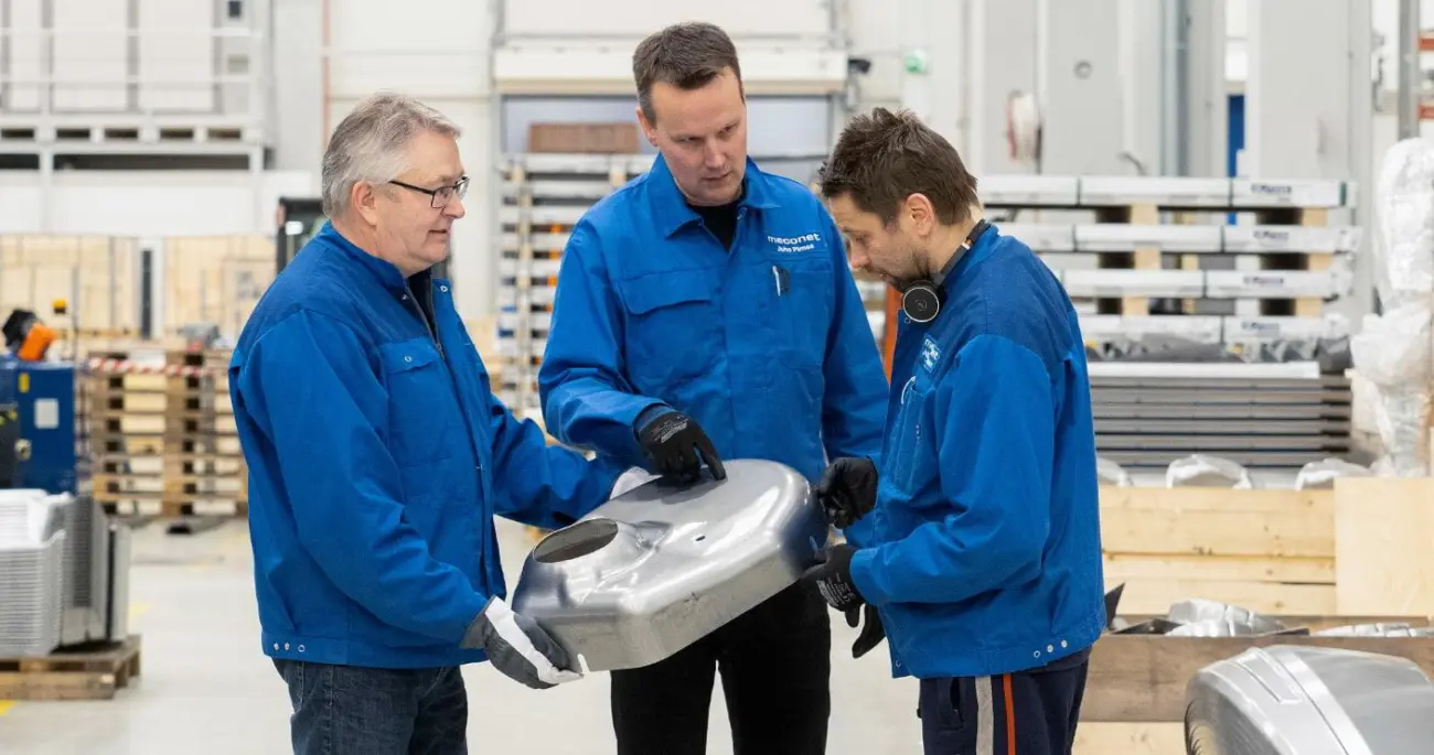 Tre medarbetare på Meconet granskar en stor formad metallkomponent i produktionslokalen.
