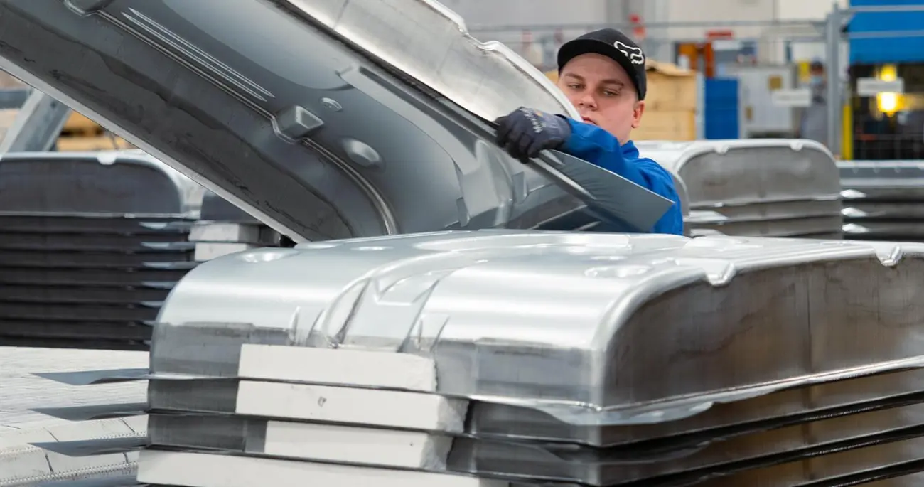 A Meconet employee handles a large formed sheet metal part beside a stack of similar parts.