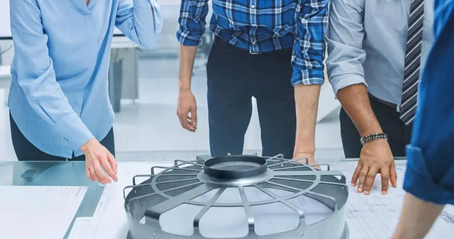 A group examines a large circular component and technical drawings on a table.