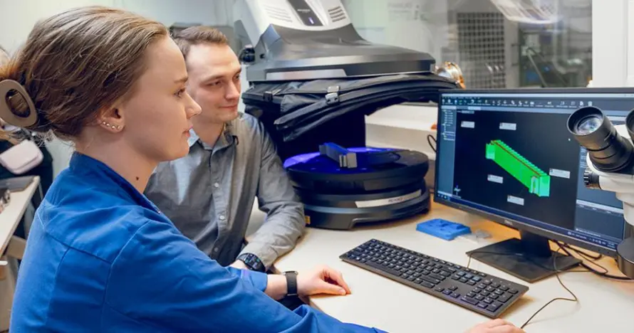 Zwei Personen betrachten ein 3D-Modell eines Bauteils auf einem Computerbildschirm neben Messgeräten.