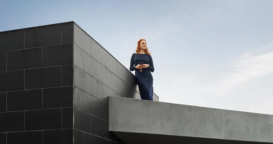 Eine Person hält ein Telefon auf der Terrasse eines modernen Gebäudes.