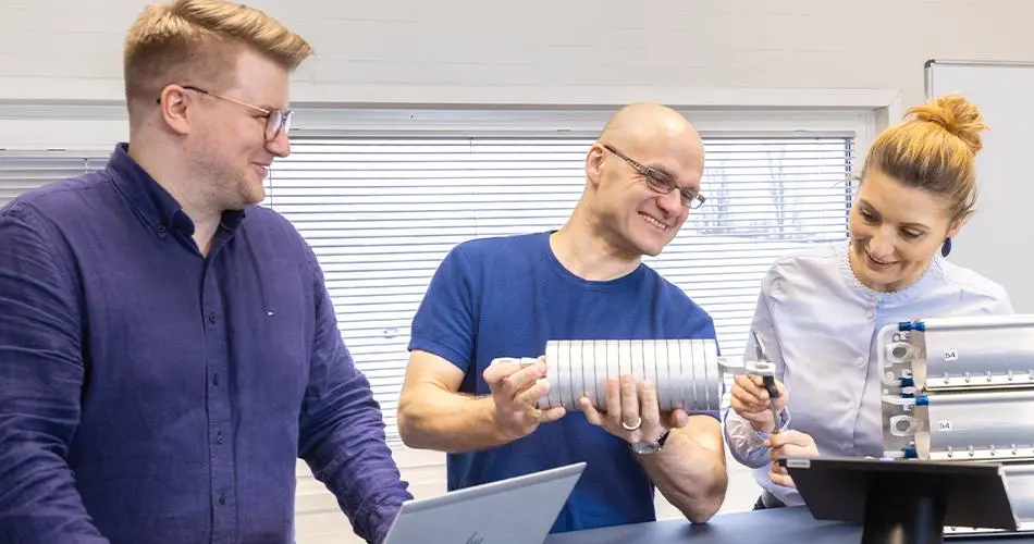 Drei Personen betrachten eine zylindrische Metallbaugruppe an einem Arbeitstisch.