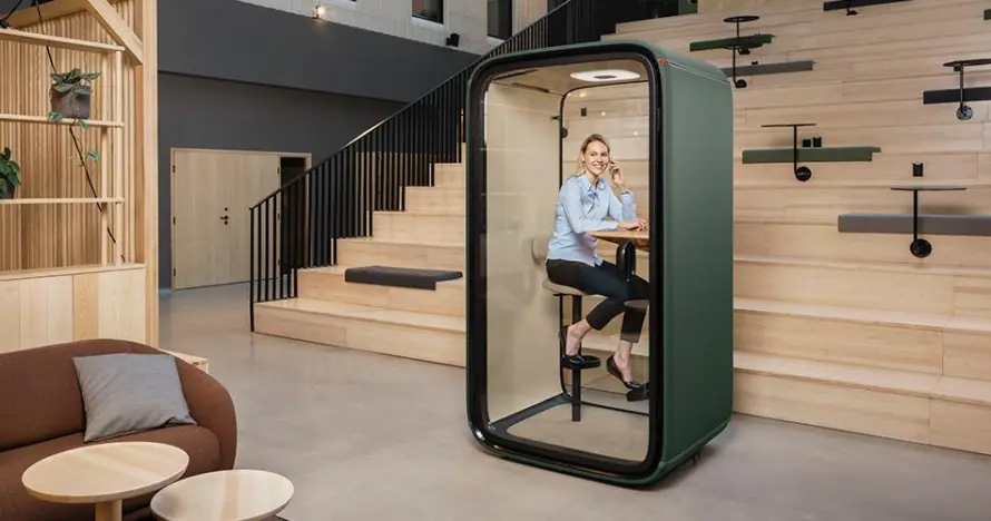 Eine Person sitzt in einer grünen Framery One-Arbeitskabine neben einer Treppe in einem offenen Bürobereich.